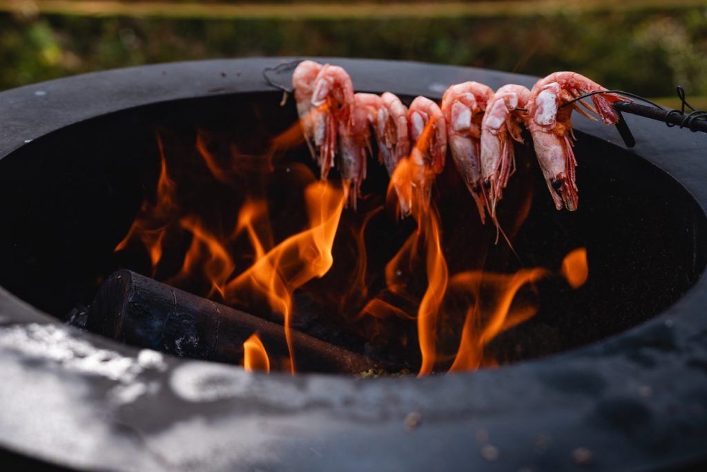 Garnalen gespiesd op een metalen staaf worden gegrild boven een open vuur op een ronde barbecuegrill buiten, terwijl de Noorwegen Nomads genieten van de verse vangst onder de gloeiende middernachtzon bij de fjorden.