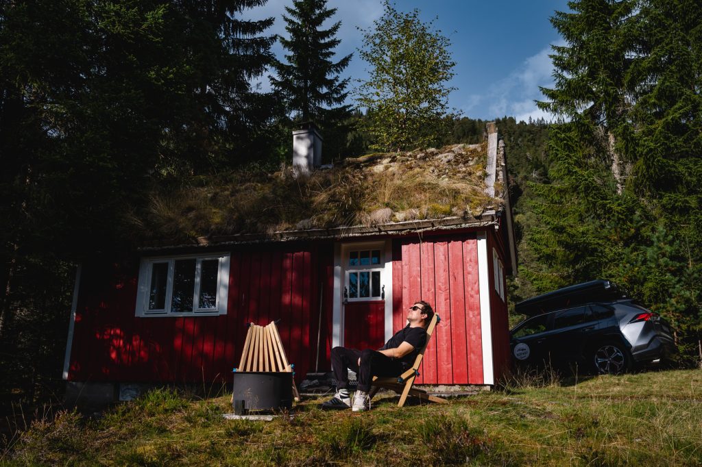 Een persoon ontspant op een stoel voor een kleine rode houten hut met een groen dak, omringd door bomen en zonlicht onder de middernachtzon, met fjorden in de buurt en een auto dichtbij geparkeerd - perfect voor Noorwegen Nomads.