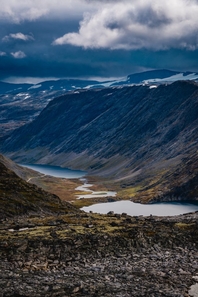 Een dramatisch berglandschap met donkere, steile kliffen, verspreide sneeuwvlokken en een kronkelend fjord onder een bewolkte, stemmige lucht. De rotsachtige voorgrond en gelaagde heuvels roepen de wilde schoonheid op die Noorwegen Nomads omarmt.