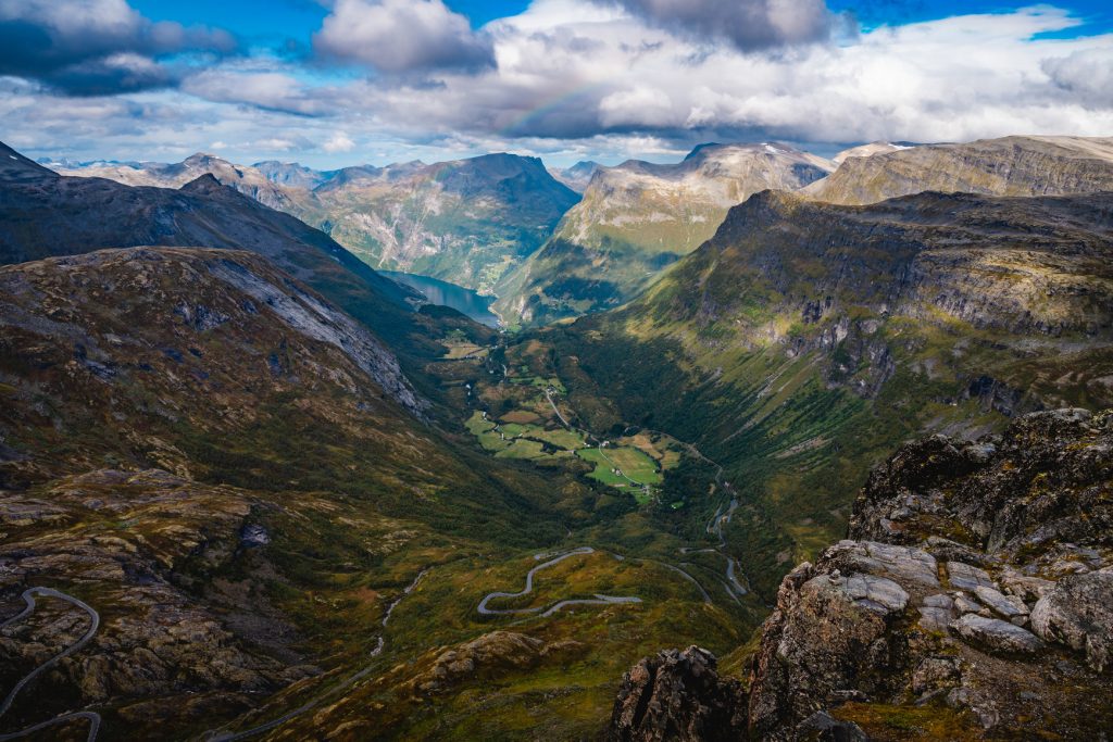 Een weids uitzicht op een weelderige groene vallei omringd door steile, rotsachtige bergen onder een gedeeltelijk bewolkte hemel, met een kronkelende weg op de bodem van de vallei en een fjordenmeer in de verte genesteld tussen de pieken - een perfect tafereel voor Noorwegen Nomads die de middernachtzon achtervolgen.