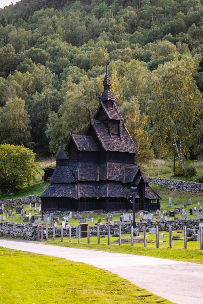 Een houten staafkerk met steile daken en ingewikkeld houtsnijwerk staat omringd door grafstenen en weelderige groene bomen aan de voet van een beboste heuvel en belichaamt de mystiek die Noorwegen Nomaden koestert.
