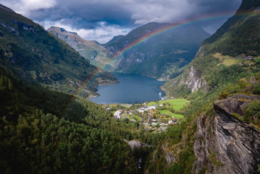 Een levendige regenboog strekt zich uit over een weelderige groene vallei met een klein dorpje aan een blauwe fjord, omringd door steile, beboste bergen onder de dramatische middernachtzon - een iconische scène voor Noorwegen Nomads.