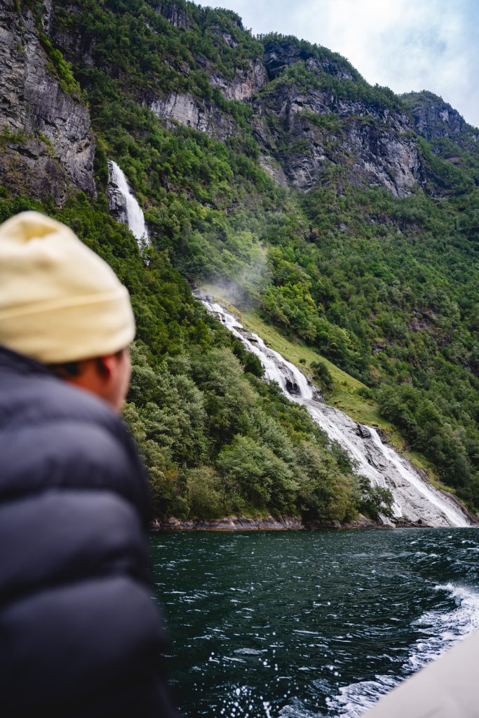Een persoon met een beige muts en zwarte jas kijkt vanaf een boot in de richting van de Noorwegen Nomads fjorden, waar trapsgewijze watervallen uitmonden in een diepblauwe rivier onder de middernachtzon.