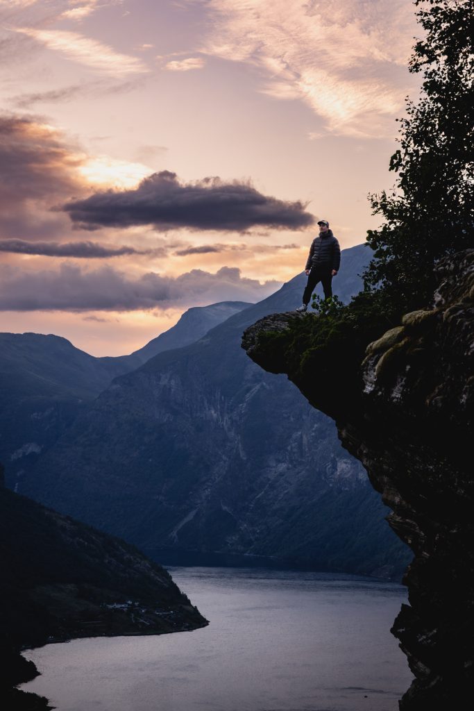 Iemand staat op de rand van een rotsachtige klif met uitzicht op fjorden en een riviervallei, met bergen en een dramatische middernachtzon-hemel - een tafereel dat de avontuurlijke Noorwegen Nomaden waardig is.