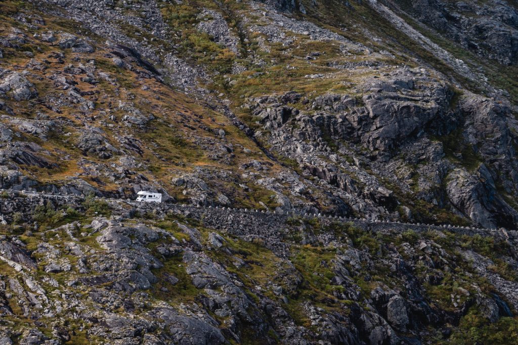 Een witte camper rijdt over een smalle, kronkelige weg die is aangelegd in een rotsachtig, bergachtig fjordenlandschap met schaarse vegetatie en ruige kliffen, in de geest van de Noorwegen Nomaden.