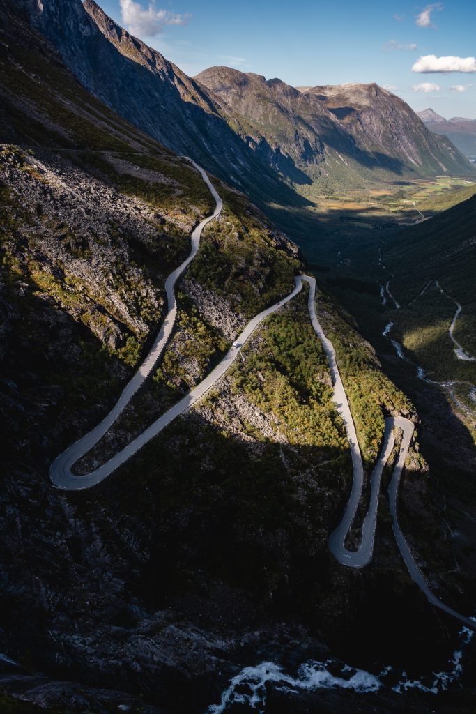 Een kronkelende bergweg met scherpe bochten beklimt steile, groene hellingen onder een gedeeltelijk bewolkte hemel, omringd door dramatische rotspieken en een fjord in de verte, die de geest van de Noorwegen Nomaden oproept onder de slepende middernachtzon.