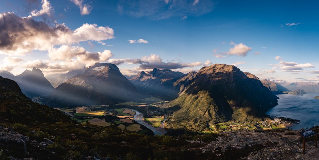 Panoramisch uitzicht op een weelderige vallei omringd door ruige bergen, met zonlicht dat door wolken in het landschap schijnt en een rivier die naar de fjorden kronkelt - een betoverend tafereel dat de geest van de Noorwegen Nomaden oproept.