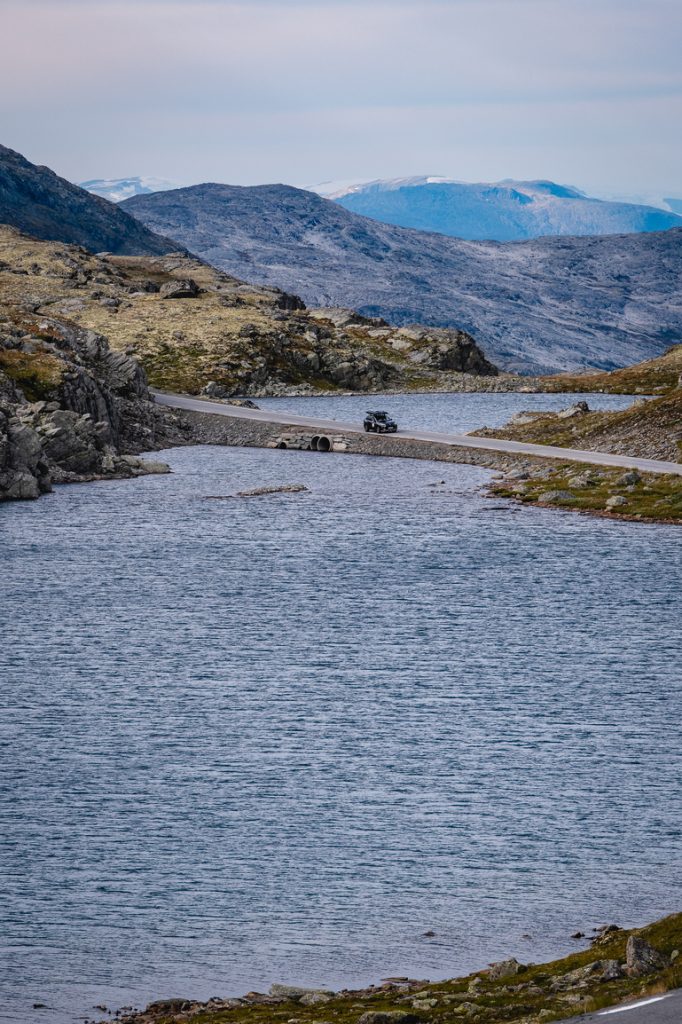 Een auto rijdt over een kronkelige weg langs een groot bergmeer in fjordenland, omringd door rotsachtige heuvels en bergen in de verte onder de beroemde middernachtzon van de Noorwegen Nomads.