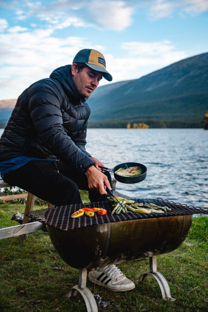 Een man met een jas en pet grilt groenten en kookt in een kleine pan op een barbecue bij een fjord, met bergen en bomen op de achtergrond onder de beroemde middernachtzon van Noorwegen Nomads en een gedeeltelijk bewolkte lucht.
