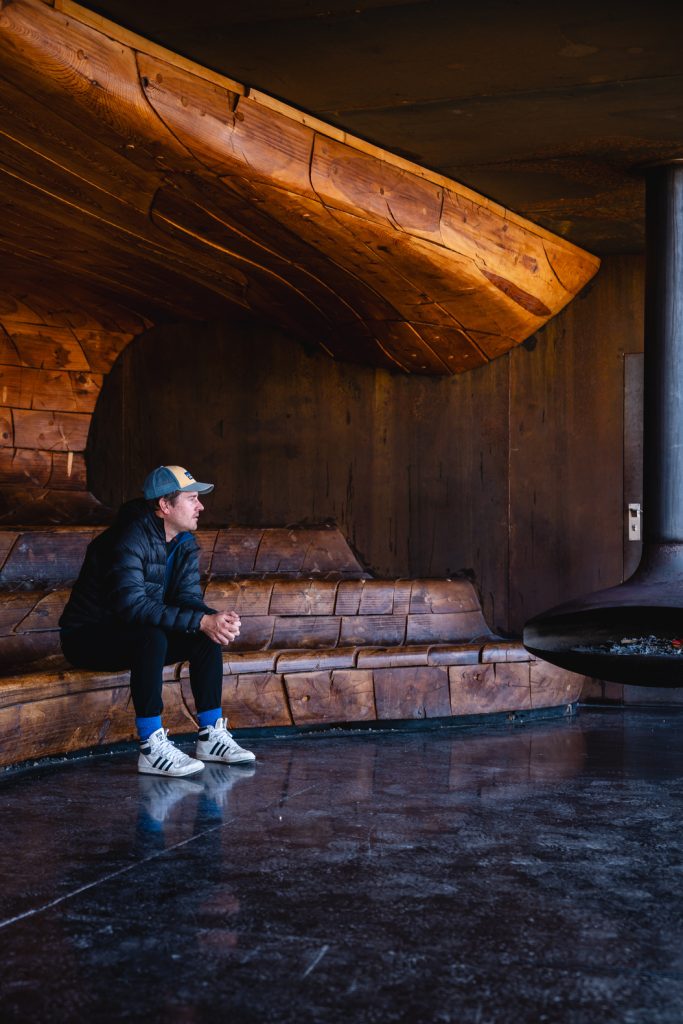 Een man met een grijze pet, zwarte jas en Adidas sneakers zit op een gebogen houten bank in een moderne kamer met houten muren en een hangende open haard, die de gezellige sfeer van Noorwegen Nomads onder de middernachtzon oproept.