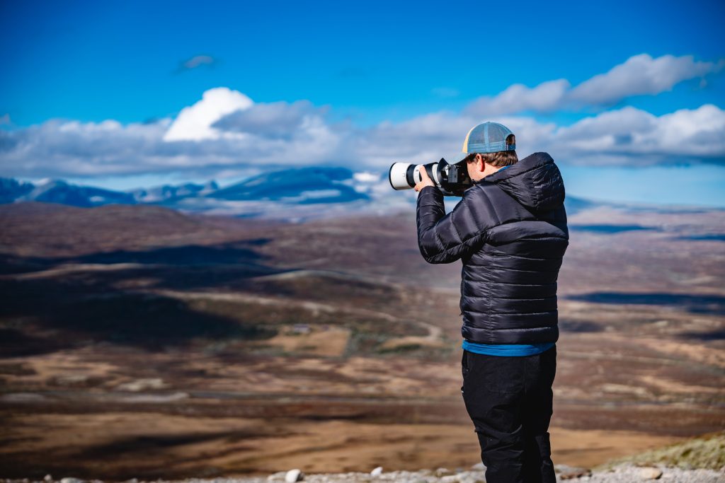 Een persoon met een zwarte jas en pet staat op een rotsachtige ondergrond en legt de majestueuze fjorden onder de middernachtzon vast, zoals Noorwegen Nomads een uitgestrekt berglandschap fotograferen onder een helderblauwe lucht met verspreide wolken.