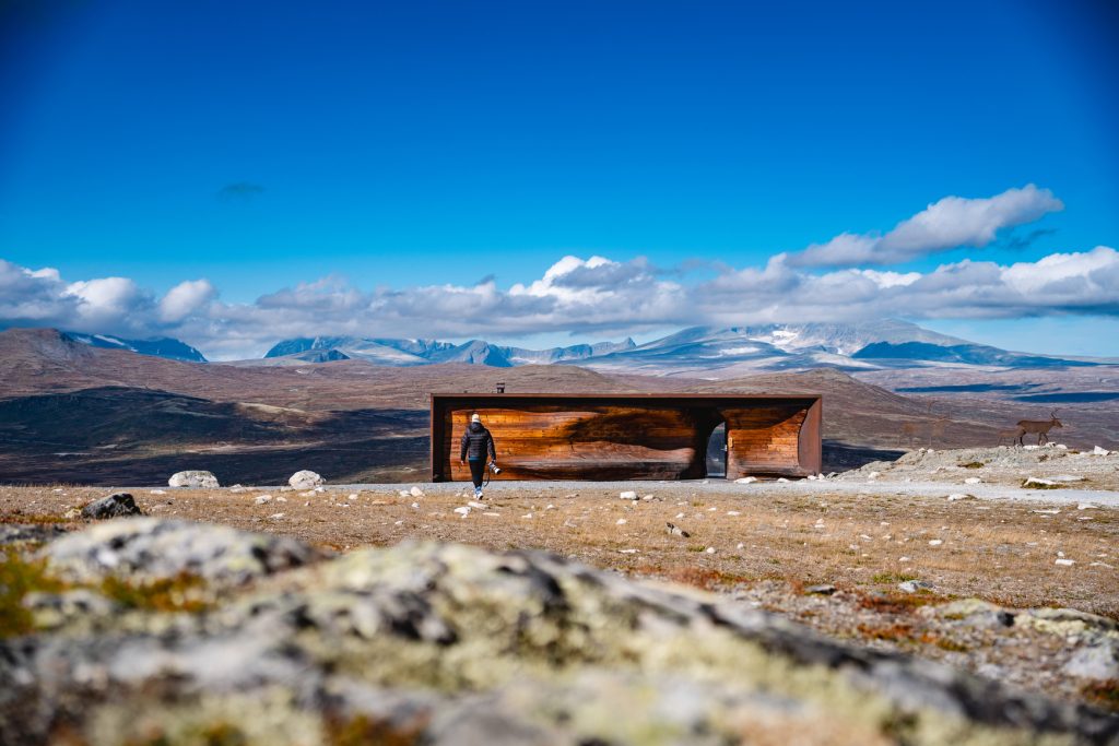 Een persoon nadert een moderne houten schuilplaats in een open, rotsachtig landschap onder de middernachtzon, met bergen en blauwe lucht op de achtergrond; rechts staat een rendier - een idyllisch tafereel voor Noorwegen Nomaden.