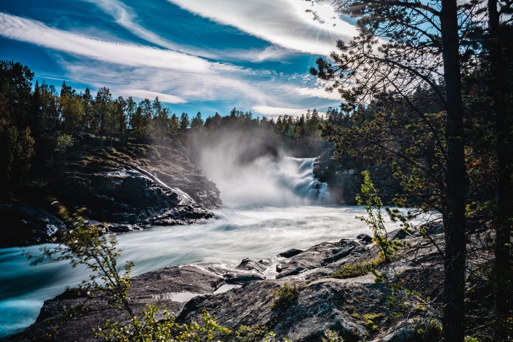 Een krachtige waterval stort zich uit over rotsachtig terrein in fjordenland, omringd door pijnbomen en mist. Dramatische, gestreepte wolken vullen een helderblauwe hemel terwijl het zonlicht van de middernachtzon er doorheen schijnt en dit Noorwegen Nomads avontuur verlicht.