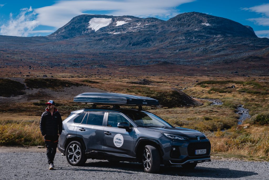 Een persoon in outdoorkleding staat naast een grijze SUV met dakkoffer, geparkeerd op een grindweg onder de middernachtzon, met fjorden en bergen die zich uitstrekken aan de horizon-Noorwegen Nomaden die de wilde schoonheid verkennen.