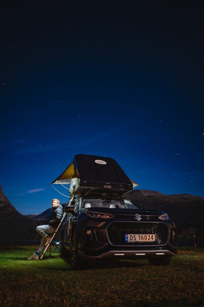 Een persoon zit in een stoel naast een zwarte SUV met een daktent die 's nachts is opgezet onder de middernachtzon. Sterren verlichten de heldere hemel, fjorden strekken zich in de verte uit en bergen omlijsten dit Noorwegen Nomads avontuur.