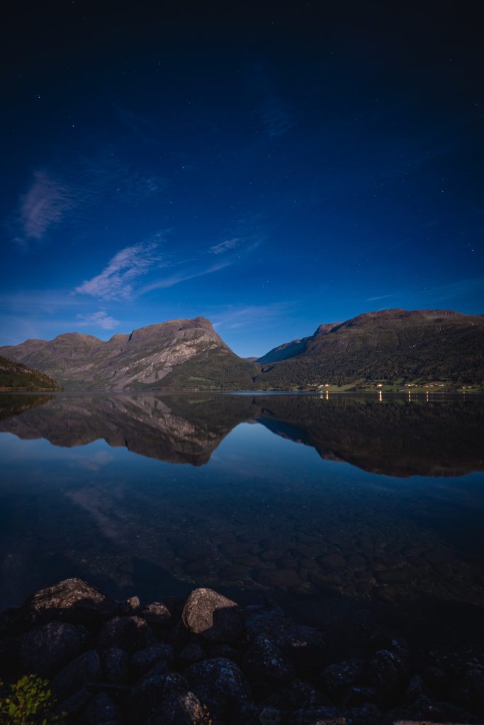 Een rustig meer in Noorwegen weerspiegelt de nabijgelegen bergen onder de middernachtzon met sterren, met uitzicht op fjorden en rotsen zichtbaar langs de kustlijn op de voorgrond.