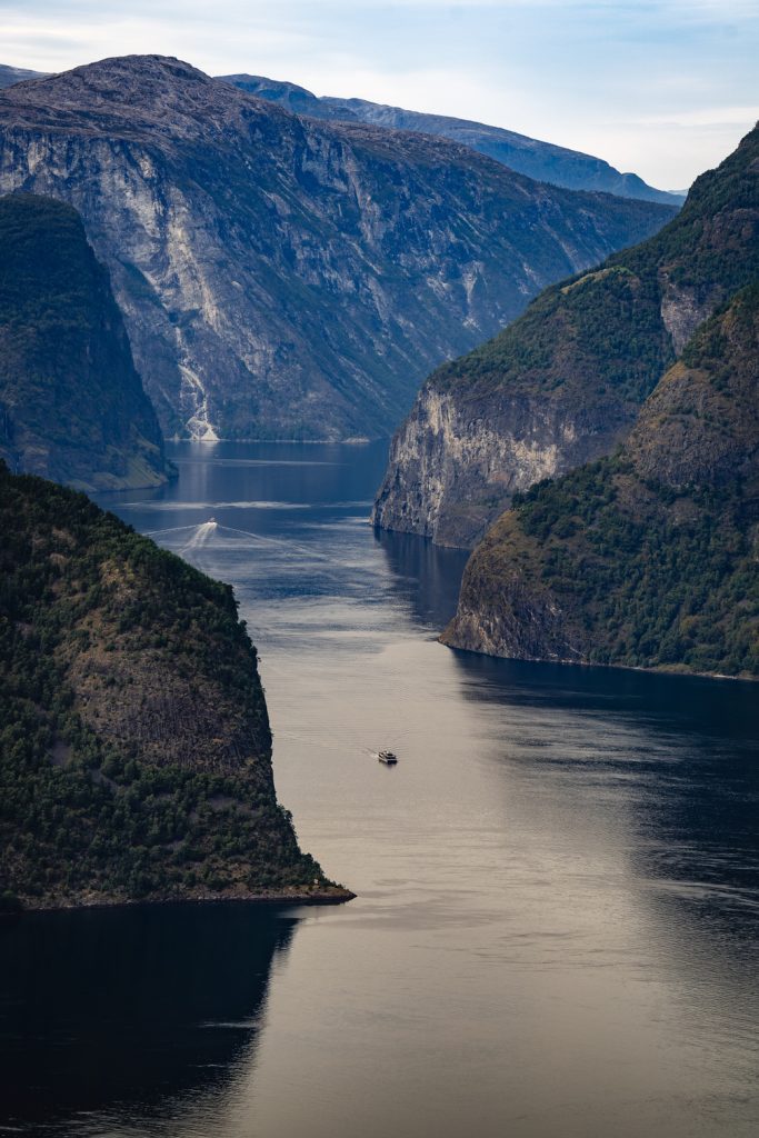 Een boot vaart door fjorden in diepblauw water, omringd door steile, beboste kliffen en rotsachtige bergen onder de betoverende middernachtzon van de Noorwegen Nomads en een gedeeltelijk bewolkte hemel.