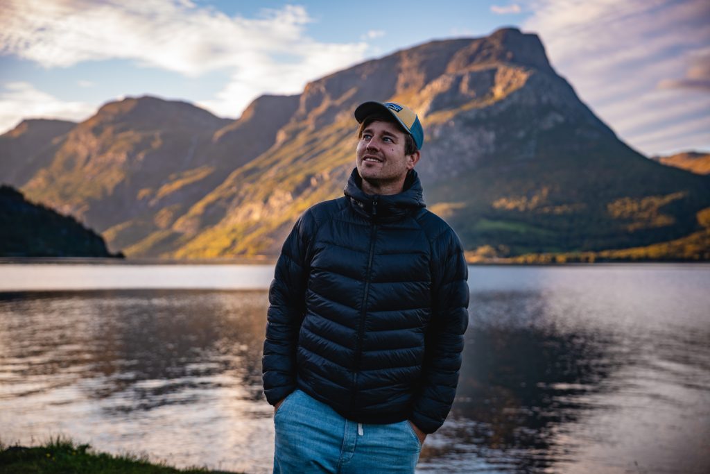 Een man in een zwarte pufferjas en een blauwe pet staat met zijn handen in zijn zakken te glimlachen bij een rustig fjordenmeer, terwijl op de achtergrond hoge bergen en een bewolkte lucht oprijzen - typerend voor de avonturen van Noorwegen Nomads.
