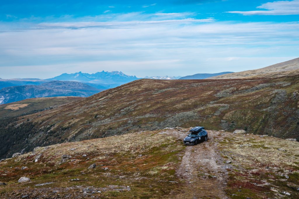 Een zwarte SUV staat geparkeerd op een rotsachtig, onverhard pad boven op een bergrug met verre bergtoppen onder de middernachtzon. Het landschap, dat doet denken aan de fjorden, bestaat uit grashellingen, rotsen en weidse uitzichten die geschikt zijn voor Noorwegen Nomaden.