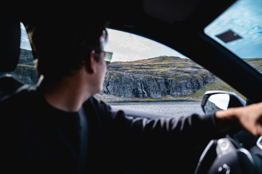 Een persoon rijdt in een auto en kijkt uit het raam naar rotsachtige heuvels en grasland onder de middernachtzon. De foto, genomen vanuit de auto, legt het prachtige fjordenlandschap buiten vast, perfect voor Noorwegen Nomads.