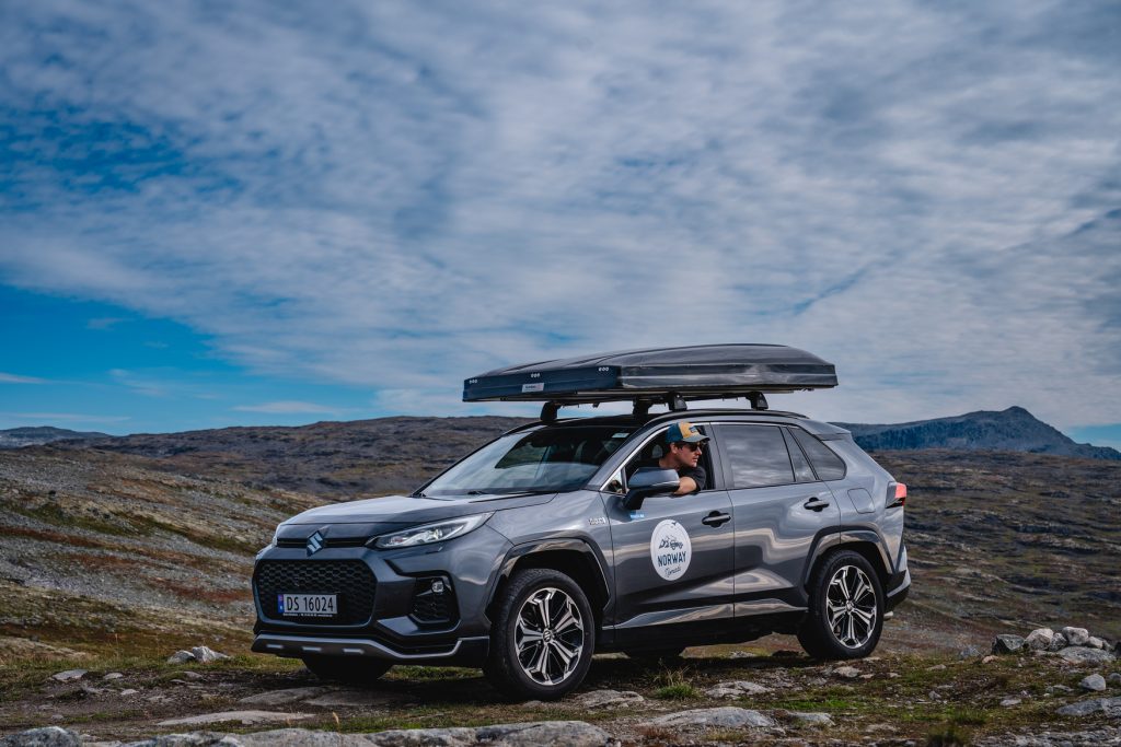 Een persoon zit in een grijze SUV met dakkoffer, geparkeerd op rotsachtig terrein onder de blauwe lucht met wolken en de verre fjorden van Noorwegen, misschien wachtend om de magie van de middernachtzon te ervaren.