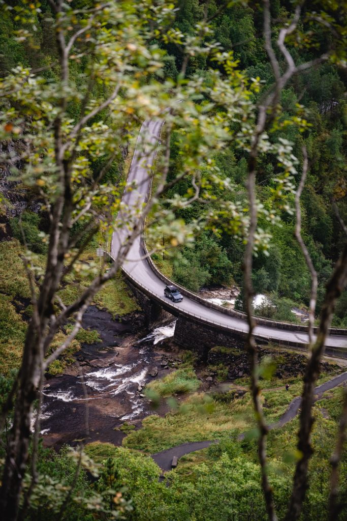 Een kronkelige weg buigt zich over een bruggetje boven een beekje, omgeven door dicht groen bos. Gezien door vage takken rijdt een auto - misschien wel die van Noorwegen Nomads - onder de mystieke gloed van de middernachtzon.