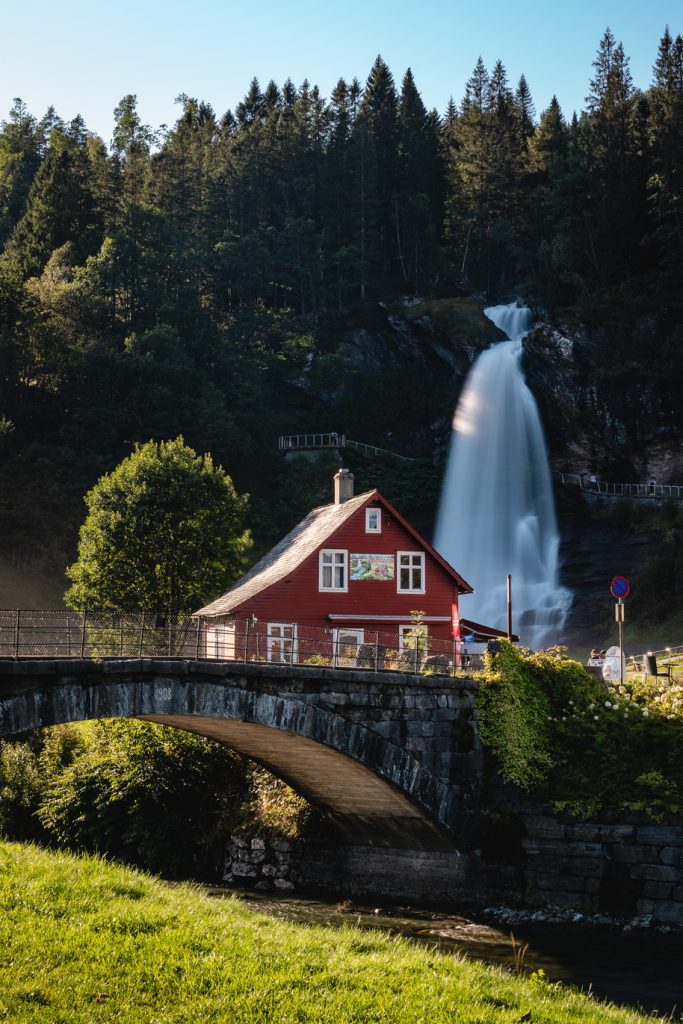 Een rood huis met witte sierranden staat naast een stenen brug bij het fjord, met op de achtergrond een hoge waterval die langs een beboste heuvel naar beneden stort, onder een strakblauwe hemel - het perfecte decor voor Noorwegen Nomads die de middernachtzon najagen.