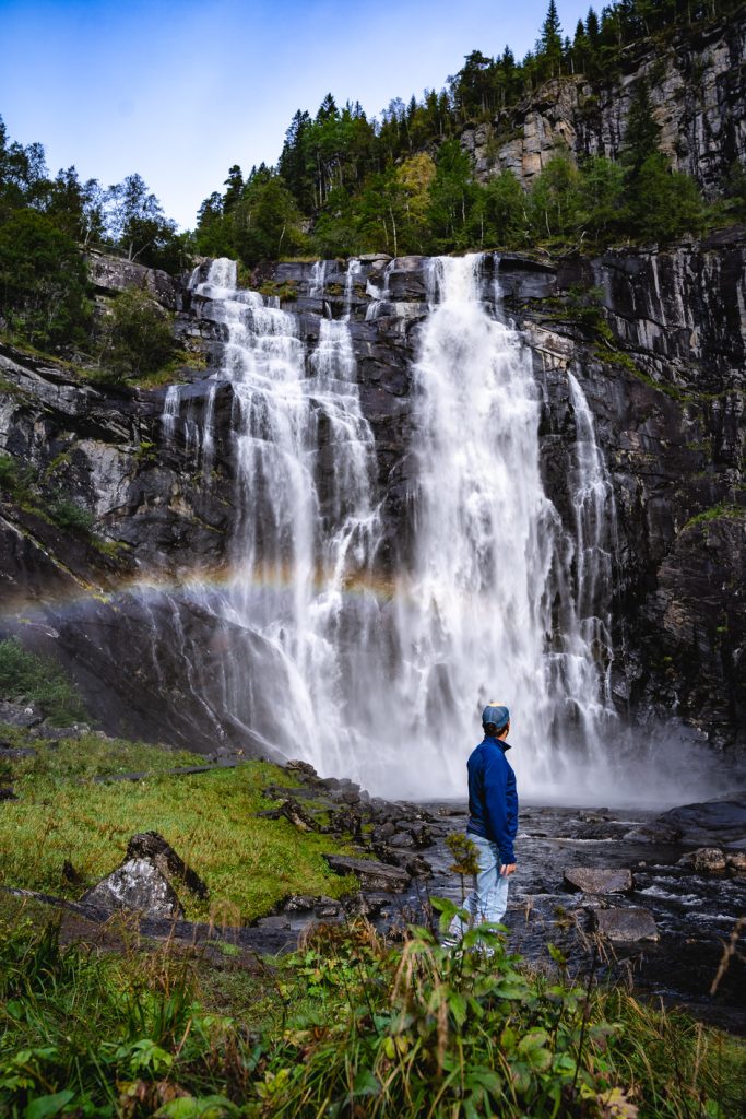 Een persoon in een blauwe jas staat op een rots en kijkt naar een hoge, brede waterval die langs een klif naar beneden stort, omringd door weelderig groen in een fjord, met een vage regenboog die glinstert aan de voet - een iconisch uitzicht voor Noorwegen Nomads.