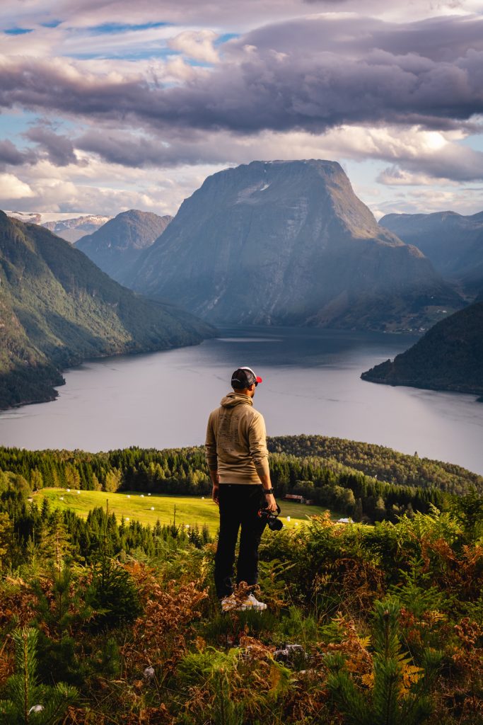 Een persoon staat op een met gras begroeide heuvel en kijkt uit over de Noorwegen Nomads fjorden, omringd door beboste bergen en dramatische wolken, met een grote, prominente piek op de achtergrond onder de gloed van de middernachtzon.