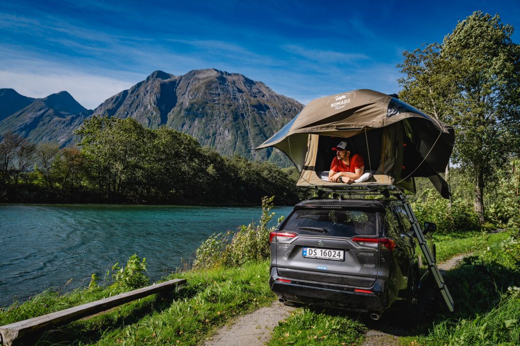 Een persoon in een rood shirt zit in een daktent op een zwarte SUV bij een rivier, omringd door groene bomen en hoge bergen, onder een strakblauwe hemel - een ideaal tafereel voor Noorwegen Nomads die de fjorden verkennen onder de middernachtzon.