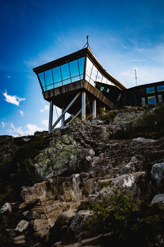Een modern gebouw met glazen wanden en een hoekig ontwerp staat op palen bovenop een rotsachtige heuvel, weerspiegelt de blauwe lucht en wolken en wordt omringd door ruig terrein en groen - een idyllisch toevluchtsoord voor Noorwegen Nomaden onder de middernachtzon in de buurt van prachtige fjorden.