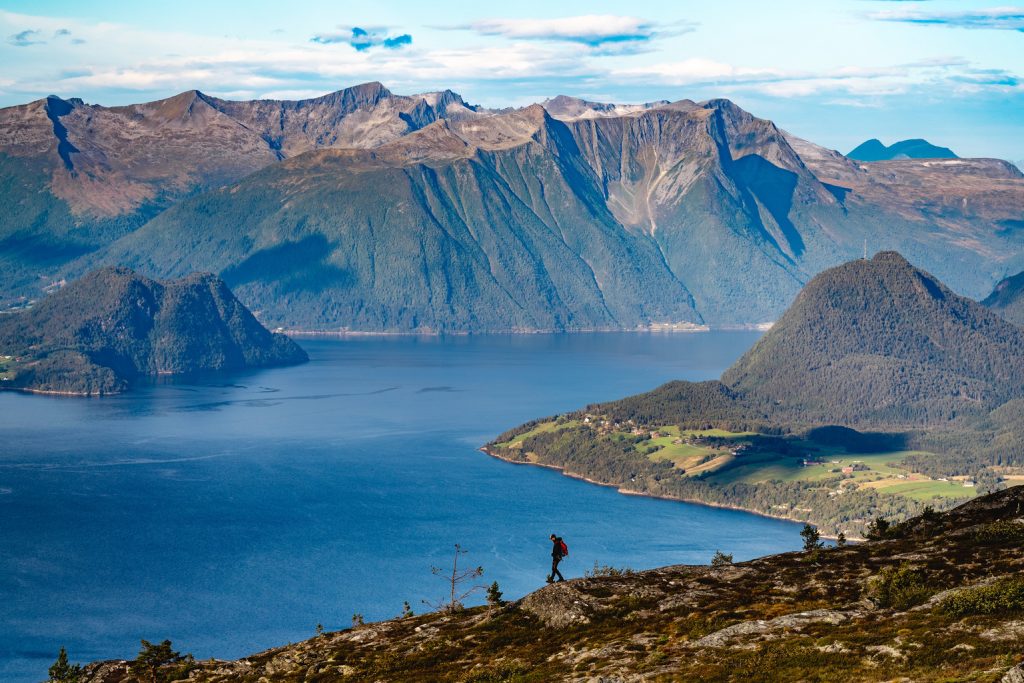 Een eenzame wandelaar loopt over een bergrug die uitkijkt over een fjord, met dramatische, steile bergen en verspreide groene valleien aan de overkant onder een gedeeltelijk bewolkte hemel - een tafereel dat perfect is voor Noorwegen Nomads.