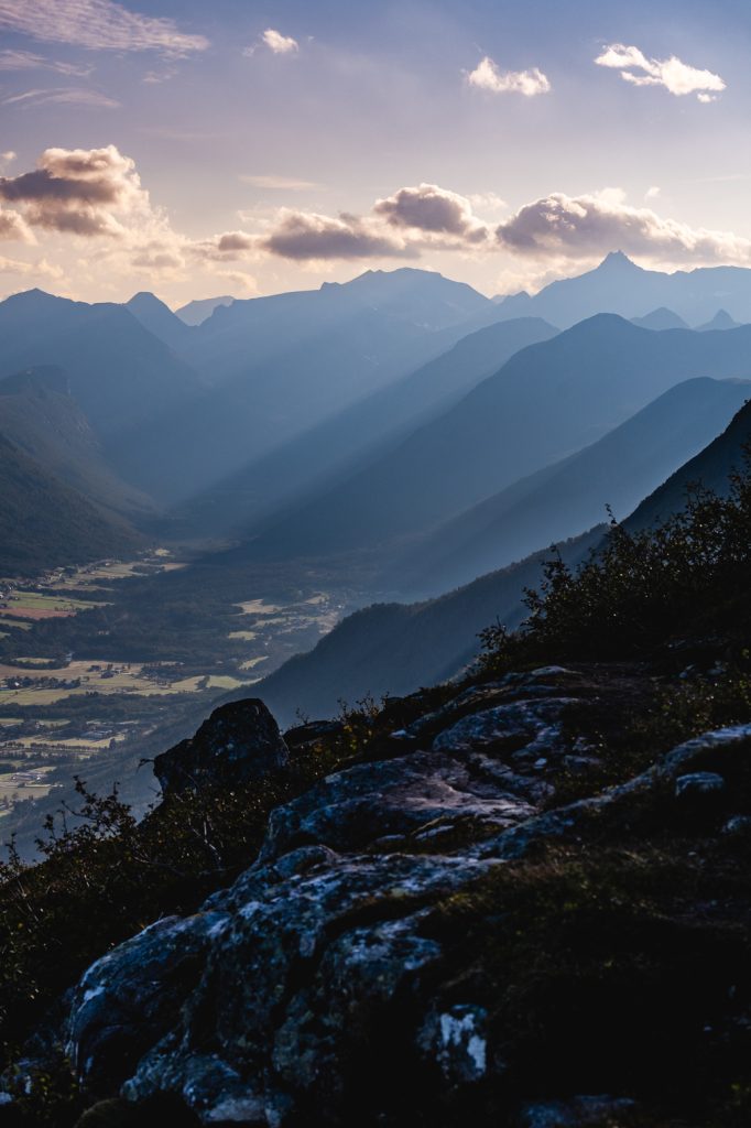 Berglandschap bij zonsondergang met gelaagde bergruggen, dramatische lichtstralen die door de wolken schijnen en een groene vallei eronder. Rotsachtige voorgrond met verspreide struiken, die de serene schoonheid van Noorwegen vastlegt onder de magie van de middernachtzon.