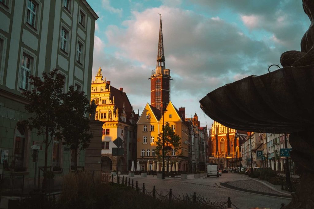 Een Europees stadsplein bij zonsondergang, dat doet denken aan Opole reizen, met kleurrijke historische gebouwen en een hoge kerktoren, met zacht zonlicht dat gevels en wolken verlicht. Een donker silhouet van een fontein staat rechts op de voorgrond.