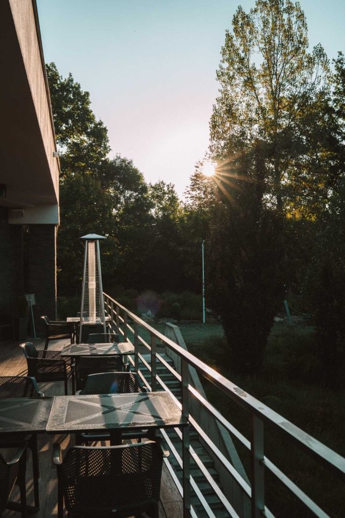 Buitenpatio met metalen tafels en stoelen naast een reling, met uitzicht op bomen en groen bij zonsondergang - een perfecte plek om te ontspannen tijdens je Opole reizen, als het zonlicht door de bomen filtert en een warme gloed werpt over deze vredige omgeving.