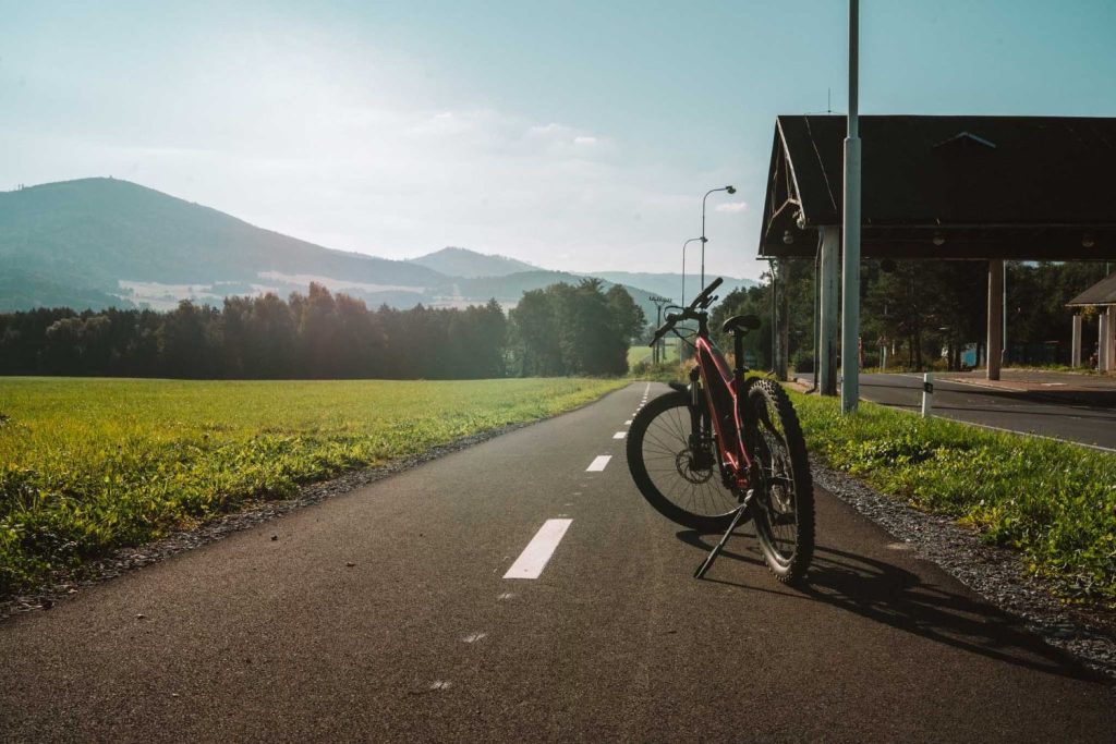 Een fiets staat op een geasfalteerd fietspad omringd door groene velden in de buurt van Opole, met bergen en bomen op de achtergrond onder een strakblauwe hemel - een perfect tafereel voor Opole reizen.