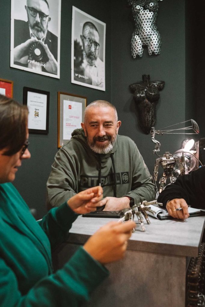 Een bebaarde man met een groene capuchon glimlacht terwijl hij praat met een vrouw met een bril aan een balie, met kunst en ingelijste foto's op de donkere muur achter hen. Ze houdt een klein voorwerp vast, wat duidt op gedeelde herinneringen aan Opole reizen tussen de metalen sculpturen op het bureau.
