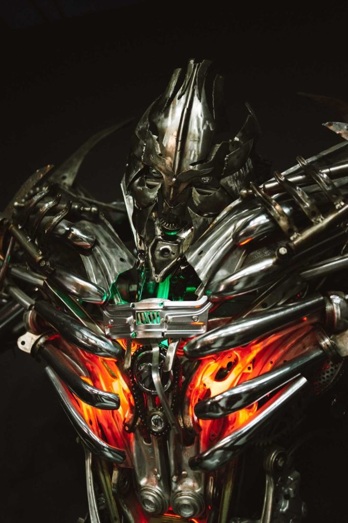 Een dreigende robotfiguur gemaakt van metalen onderdelen met oplichtende oranje en groene lampjes in zijn borst, gedetailleerd met scherpe, hoekige gelaatstrekken, wordt verlicht tegen een donkere achtergrond als een futuristische bewaker tijdens een Opole reizen avontuur.