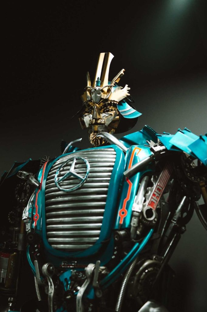 Een robot met een metalen samoerai-helm en ingewikkeld pantser, met een prominent Mercedes-Benz logo op zijn borst, staat tegen een donkere achtergrond met dramatische verlichting - een beeld dat net zo opvallend is als de avonturen van Opole reizen.