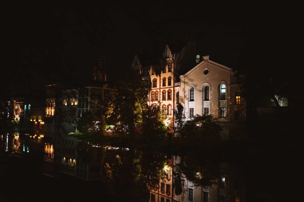 Nachtelijk uitzicht op historische gebouwen in Opole reizen verlicht door warme lichten, die weerspiegelen in een kalme rivier. De architectuur bestaat uit boogramen en ingewikkelde gevels, omringd door bomen en duisternis.