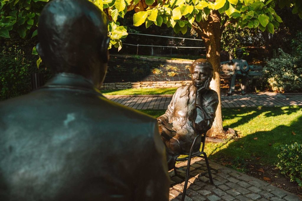 Bronzen beelden van twee mensen zitten tegenover elkaar onder een lommerrijke boom in een zonnige tuin met een stenen pad. Perfect voor een Opole reizen route, een ander standbeeld op een bankje op de achtergrond voegt diepte toe aan dit vredige tafereel.