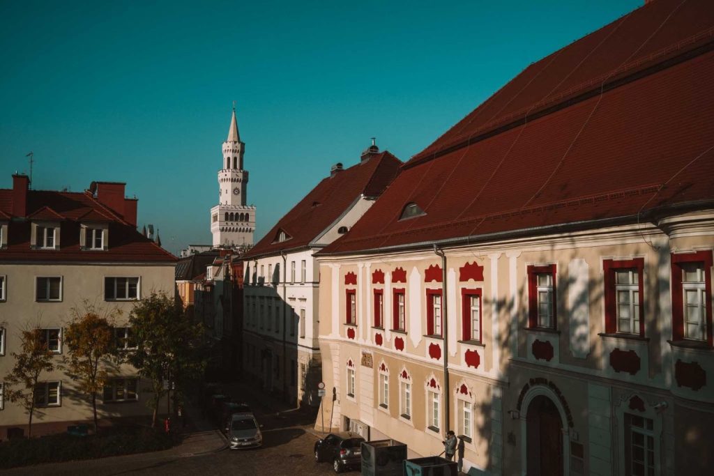 Een schilderachtige Europese straat met historische gebouwen, rode pannendaken en een hoge witte klokkentoren op de achtergrond onder een helderblauwe hemel - perfect voor wie op zoek is naar Opole reizen avonturen.