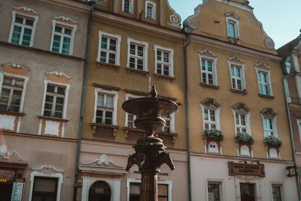 Een decoratieve metalen fontein staat voor pastelkleurige historische gebouwen met wit omrande ramen en bloembakken, onder een helderblauwe hemel - de perfecte charme voor liefhebbers van Opole reizen.