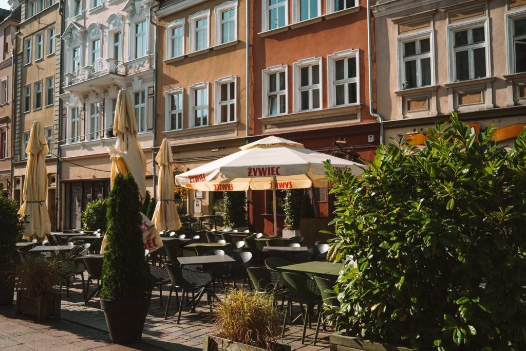 Buitencafé met lege tafels en stoelen, in de schaduw van grote crèmekleurige parasols en omringd door groene planten. Kleurrijke historische gebouwen met hoge ramen vormen de achtergrond - een perfecte plek om te ontspannen tijdens je Opole reizen op een zonnige dag.