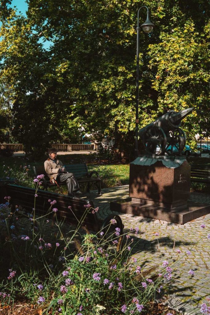 Een man zit op een bankje te lezen in een zonnig park in Opole, op reis door de geschiedenis naast een groot standbeeld van een oud kanon, omringd door groene bomen en paarse bloemen.