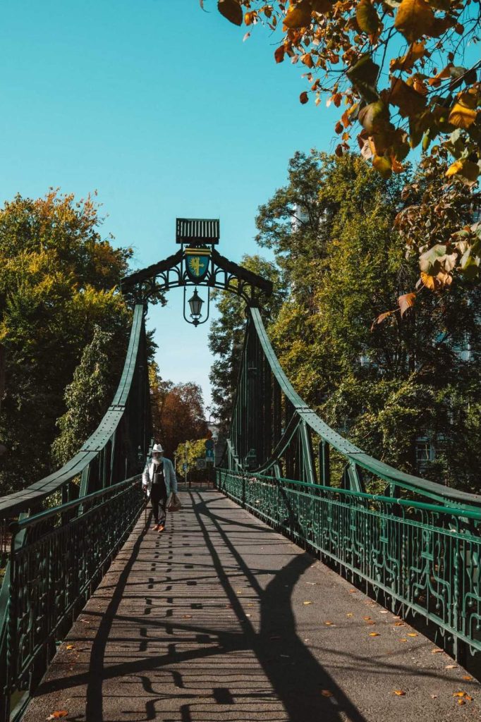 Iemand geniet van Opole reizen terwijl hij over een groene, sierlijke metalen loopbrug loopt, omringd door weelderige bomen met herfstbladeren onder een strakblauwe hemel, terwijl de schaduwen van de reling zich over het pad uitstrekken.