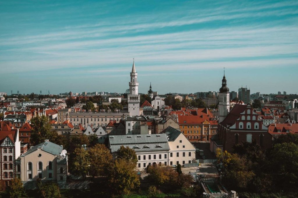 Een panoramisch uitzicht op Opole reizen onthult gebouwen met rode daken, kerktorens en weelderig groen onder een blauwe wolkenlucht. De historische architectuur en het schilderachtige stadslandschap strekken zich sierlijk uit naar de horizon.