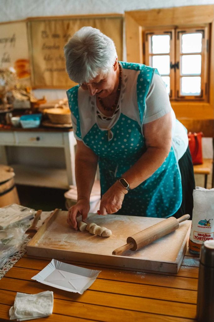 Een oudere vrouw in een blauw polka-dot schort glimlacht terwijl ze deegrolletjes klaarmaakt op een houten plank. Dit roept de warmte op van een rustieke keuken, een uitnodigend tafereel dat doet denken aan traditionele Opoolse reizen.