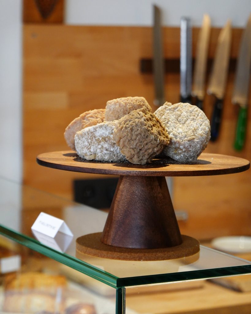 Op een houten sokkel liggen verschillende ronde stukken rustieke kaas, sommige met een grijzige korst - perfect voor een gezellige winterdate. Daarachter hangen messen aan een houten muur die Genkse Gastvrijheid oproept, terwijl een vaag kaasetiket op de glazen toonbank ligt.