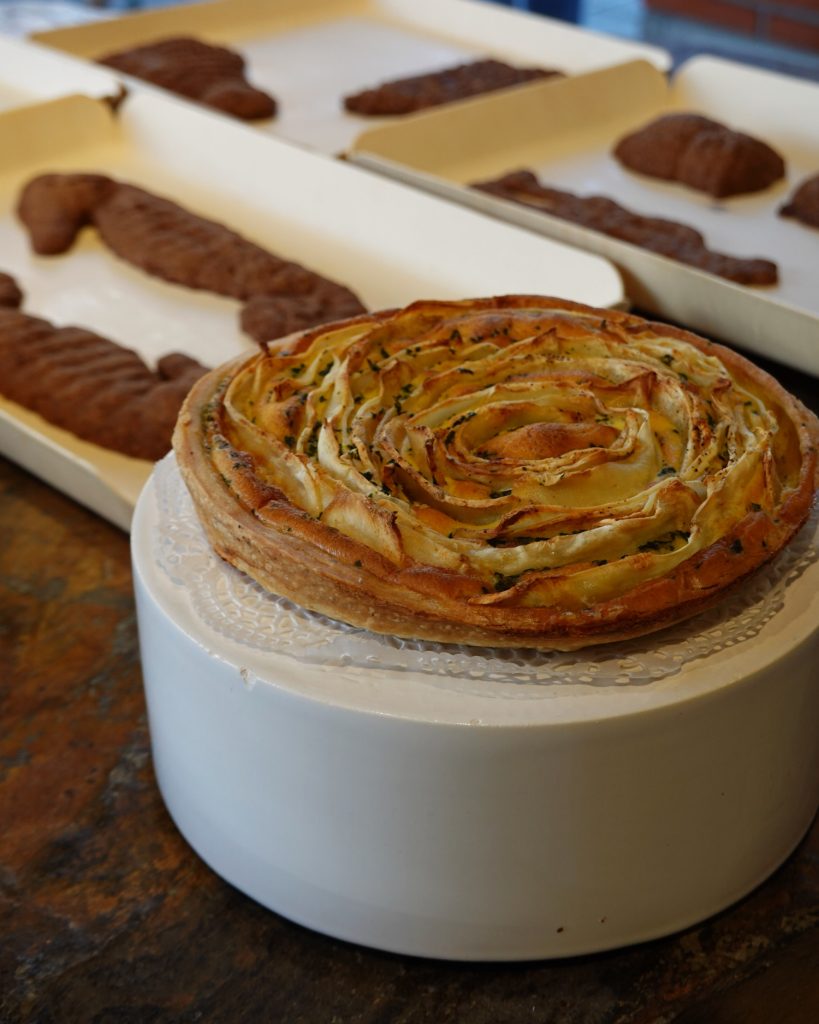 Een hartige taart met een spiraalpatroon van dun gesneden groenten staat op een witte standaard en nodigt je uit om echte Genkse Gastvrijheid te ervaren. Op de achtergrond rusten dienbladen met chocoladegebakjes op een houten oppervlak.
